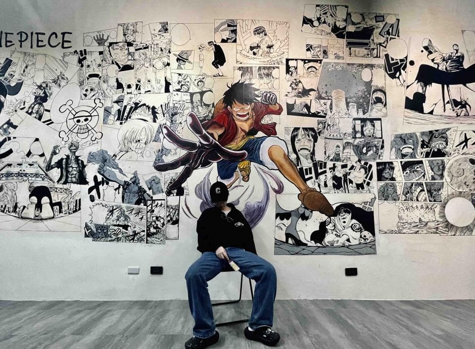 這張「海賊王」的照片是牆壁上滿滿的海賊王漫畫圖,中間有一個人坐在椅子上,身穿黑色外套和牛仔褲。