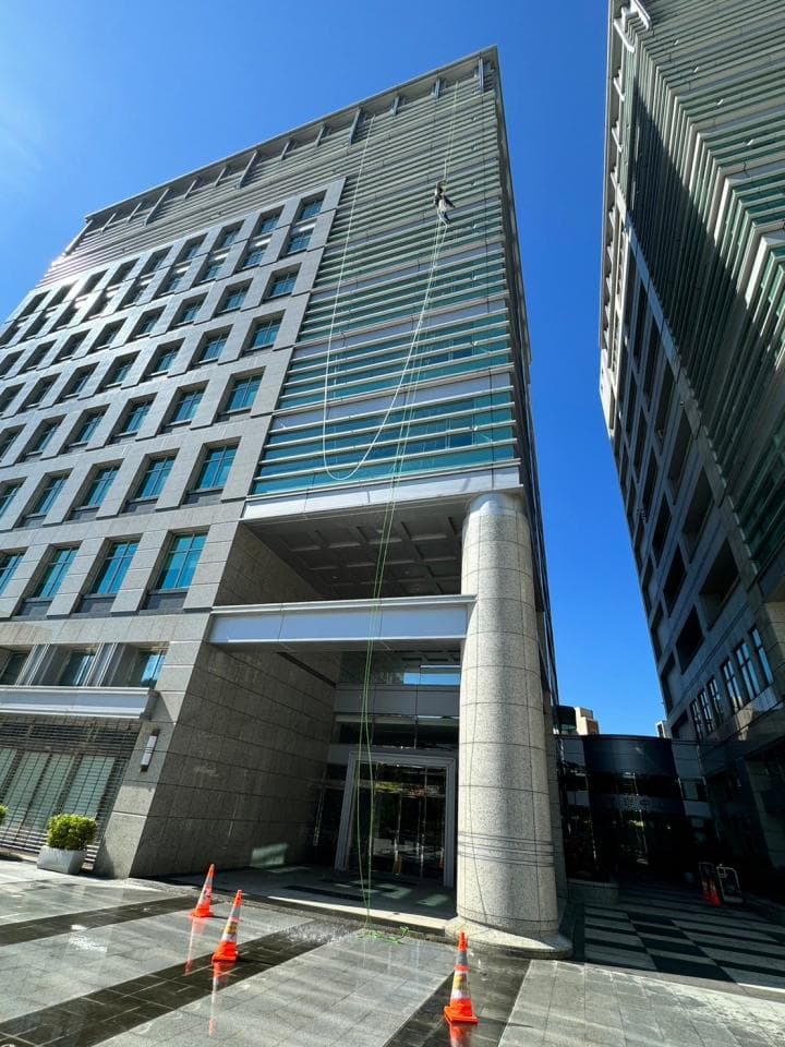 這張照片是一棟高樓大廈,照片中最重要的部分是建築物外牆上的一名工人,他正在清洗窗戶。他繫著繩索,從高處垂降下來,安全帽和工作服十分專業。