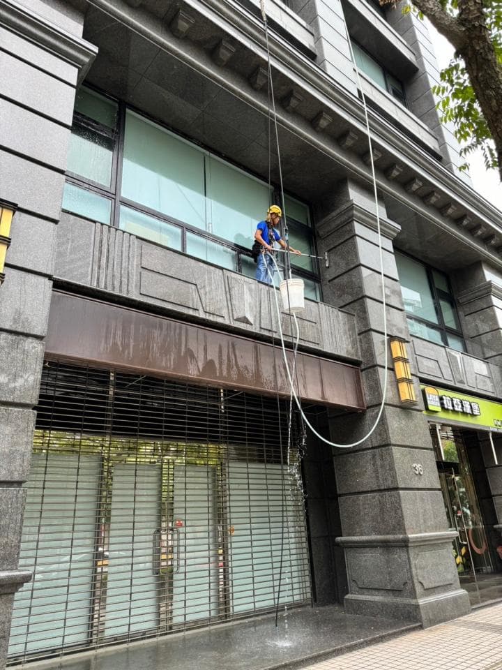 1) 照片顯示一位身穿藍色上衣和黃色安全帽的工人,他正懸吊在高樓外牆上清洗窗戶