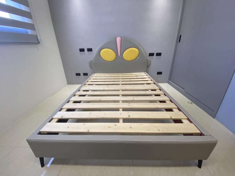 這張照片是一張有可愛造型床頭的床,床頭的形狀像一個卡通人物,床頭板是灰色,上面有兩個黃色的圓形和一個粉紅色的三角形,床墊是木質的,床架是灰色的。