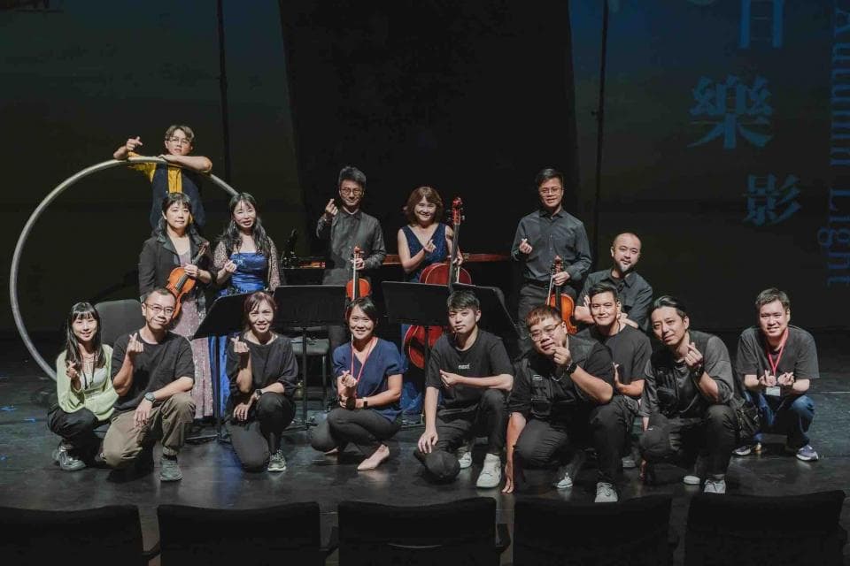 這張「一群人站在舞臺上」的照片是,一群人站在舞臺上,他們看起來像是音樂家,有人拿著小提琴,有人在唱歌,他們穿著不同的衣服,其中有幾個人戴著眼鏡。