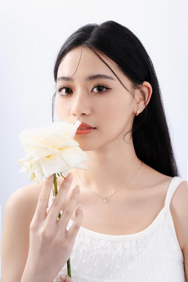 這張照片是一個年輕女子手持一朵白色玫瑰,她身穿白色背心,並戴著一條細緻的銀色項鍊。她的神情優雅而溫柔,展現出清純的氣質。