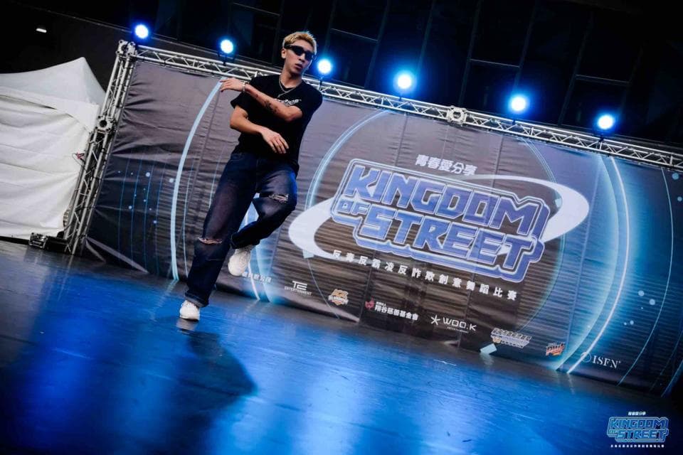 照片中有一位年輕男子,身穿黑色上衣和藍色牛仔褲,正在舞臺上表演街舞