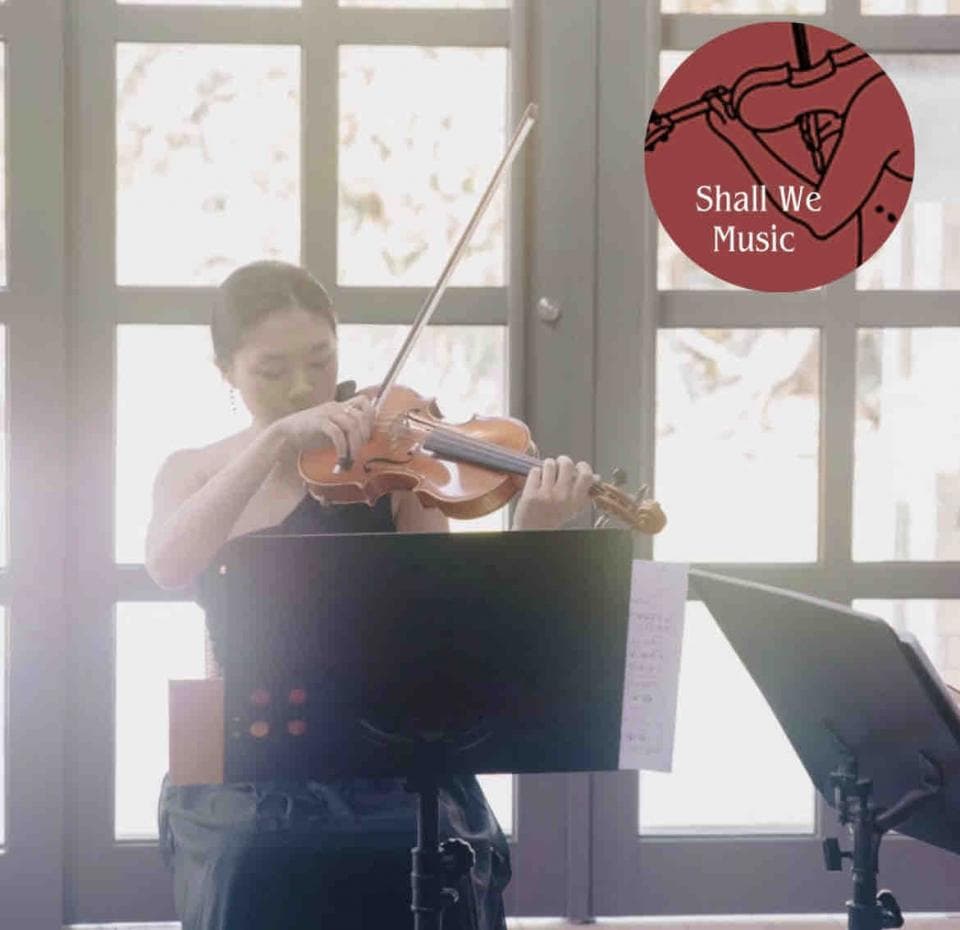 這張「一位女性在演奏小提琴」的照片是,她穿着黑色連衣裙,站在一個音樂架前,演奏着一把小提琴。她專注地演奏,背景是明亮的窗戶。