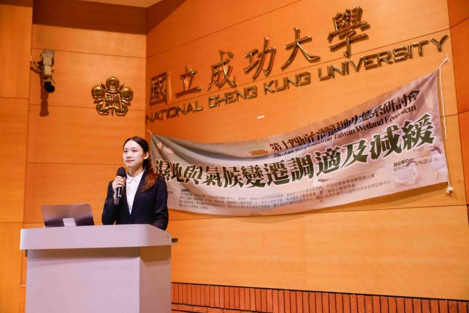這張「國立成功大學」的照片是一個女性在臺上發表演講,她拿著麥克風,身穿西裝外套。牆上掛著標語,寫著「濕地保育與永續發展」。