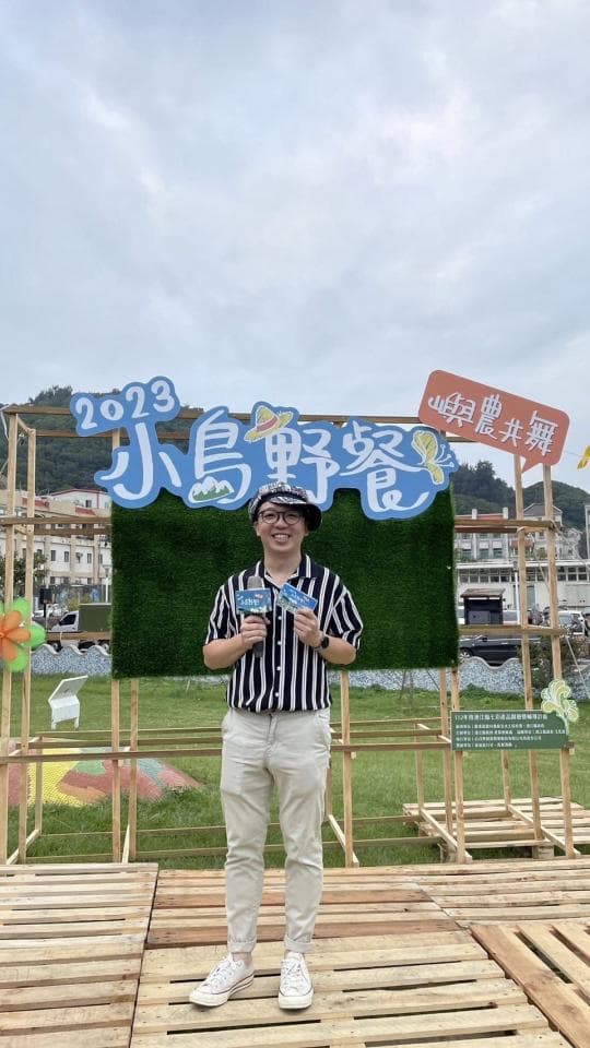 這張「小島時光」的照片是一個站在木板路上的年輕男性,他穿著黑白條紋襯衫和白色褲子,手裡拿著兩張卡片,背景是一塊綠色的草坪和一個寫著「小島時光」的藍色招牌。