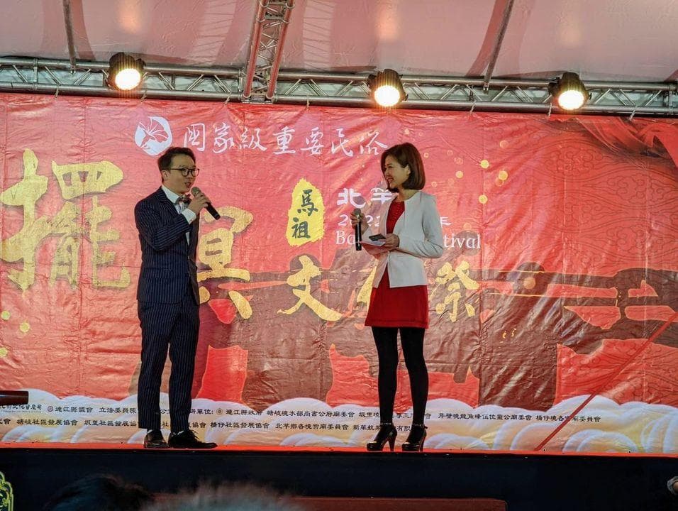 這張照片是兩位主持人站在舞台上,他們正在進行活動,其中一位主持人穿著深色西裝,手持麥克風,另一位主持人穿著白色外套,紅色裙子,手持話筒。