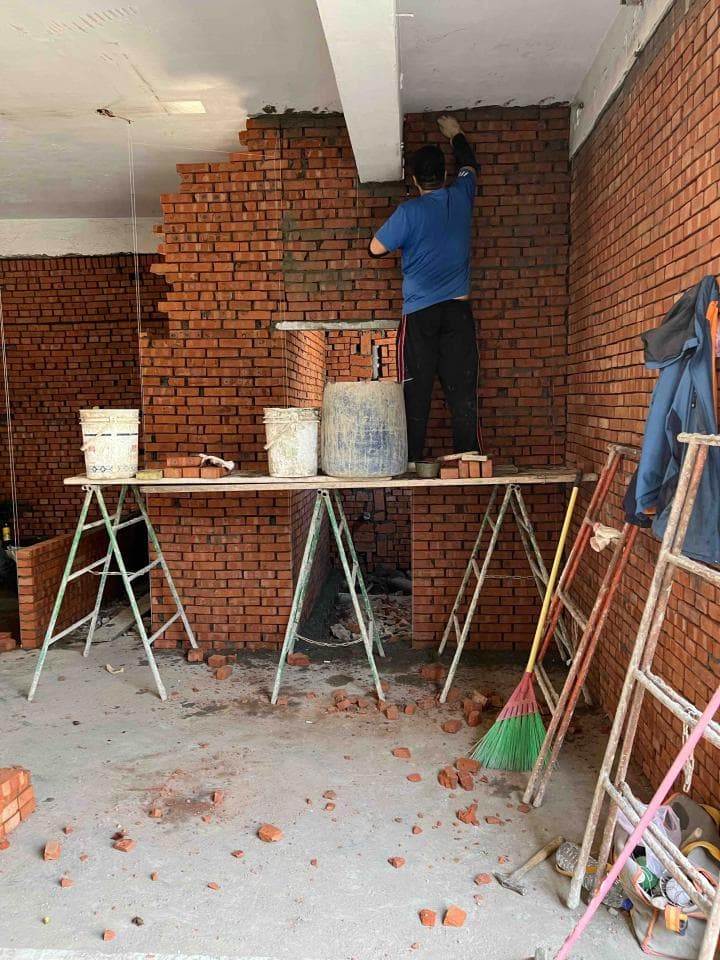 1) 照片顯示一個室內建築工地,磚牆已部分砌好,牆上有工人正在進行抹灰工程,工地上有梯子、工具和一些磚塊