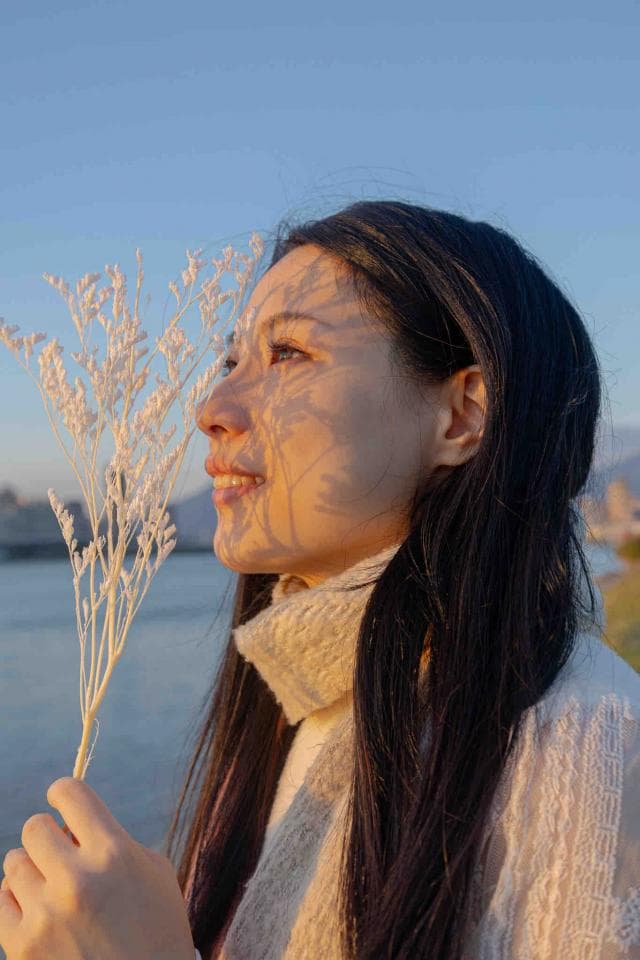 這張照片是一名年輕女子站在陽光下,她手中拿著一束白色的小花,輕輕地嗅著花香。她的表情充滿著寧靜和幸福,陽光照在她臉上,形成美麗的光影,讓人感覺溫暖和美好。