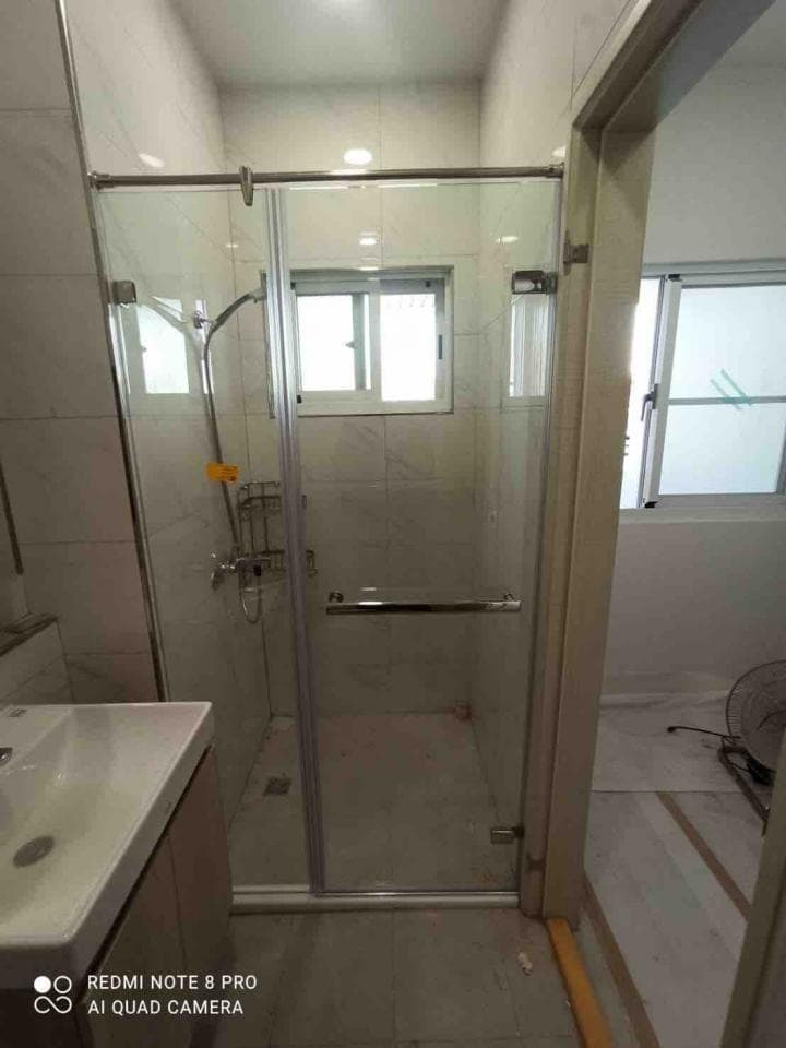 1) 照片顯示一個帶有玻璃門的淋浴間,淋浴間裡有一扇窗戶,地面上鋪著白色瓷磚,淋浴間的牆壁也是白色的瓷磚,淋浴間的門是銀色的,淋浴間外有一個水槽