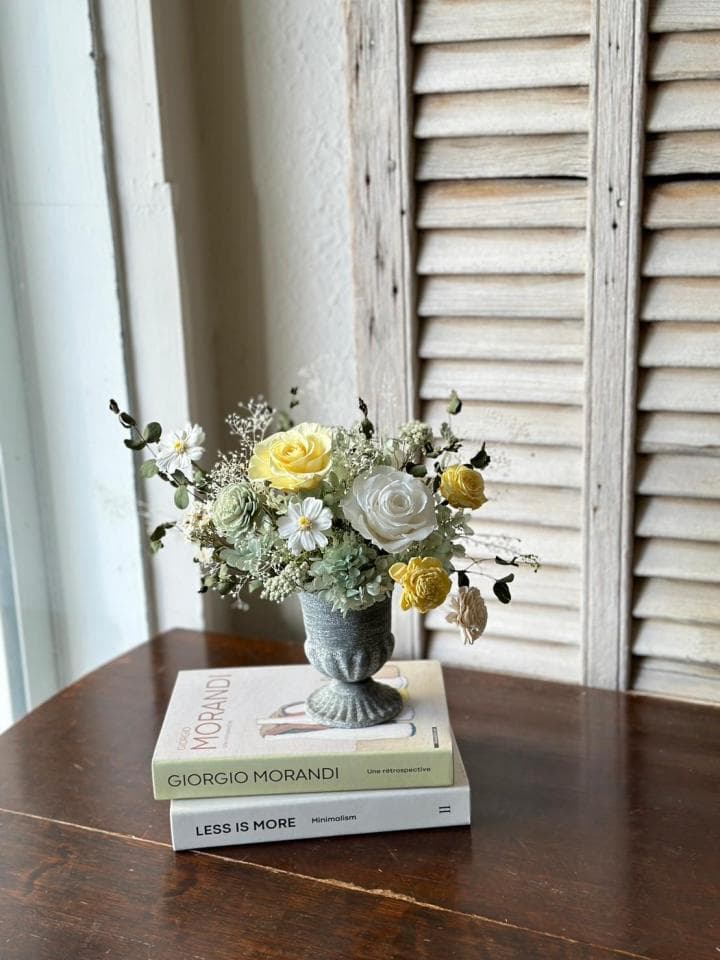 1) 照片顯示一束黃白色花朵,放置於灰色花瓶中,花瓶置於兩本書籍上,書籍擺放在木質桌面上,桌旁有白色的百葉窗