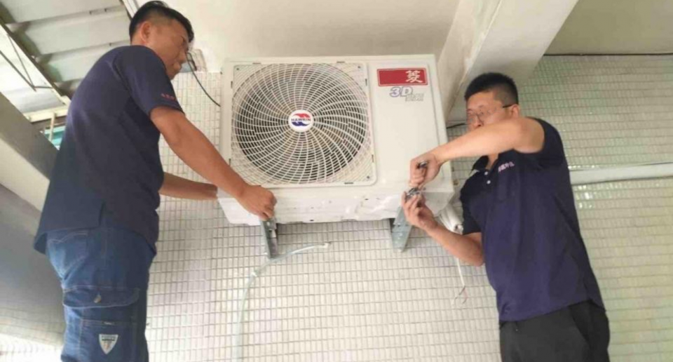 這張照片是兩位師傅正在修理冷氣。其中一位師傅站在梯子上,另一位師傅則站在地上,他們正在一起檢查和修理冷氣機。