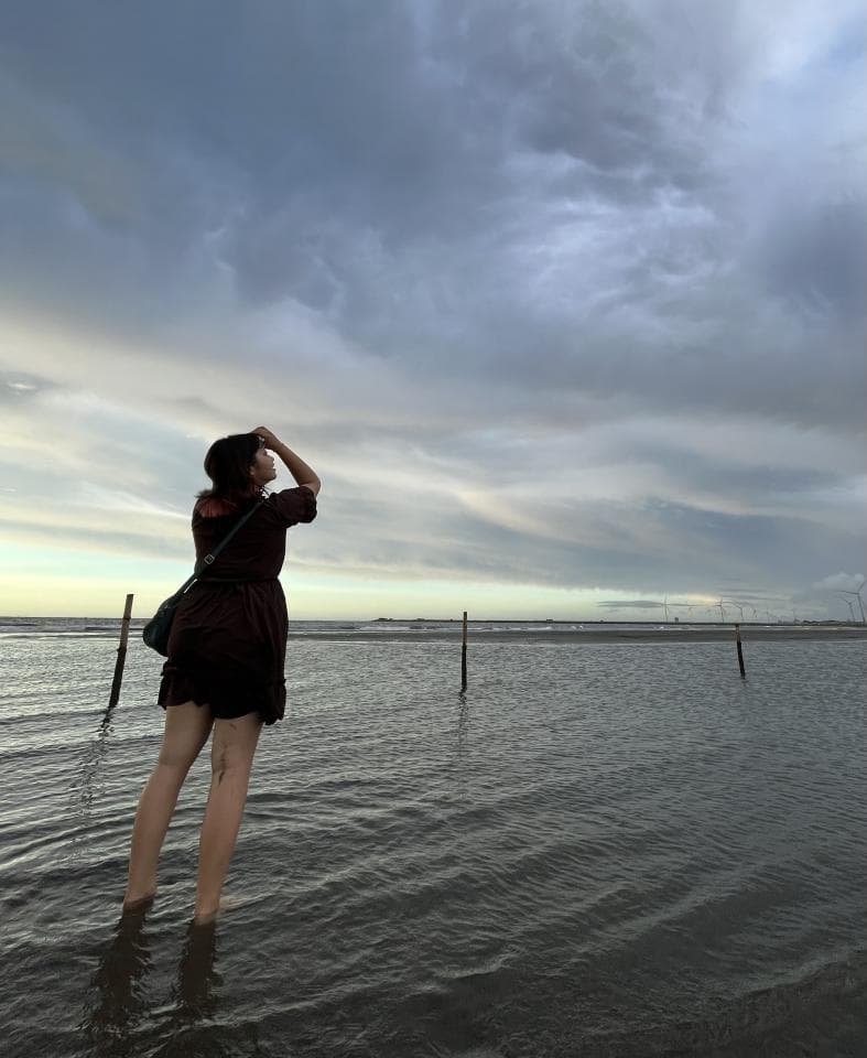 這張「海邊」的照片是一個女生站在淺灘上,她穿著黑色的洋裝,抬頭看著天空。背景是陰天,海面上泛著漣漪,遠處有幾根木樁。