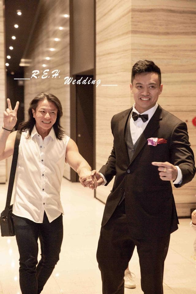 這張照片是兩個男人在婚禮上,穿著西裝的新郎牽著穿著白襯衫的伴郎的手,準備走入禮堂。