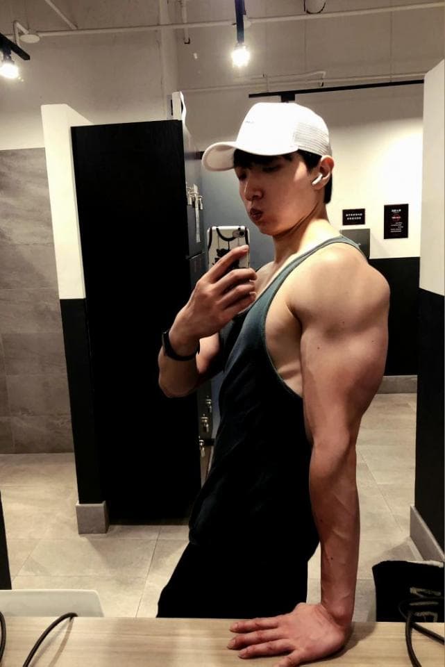 這張「健身房自拍」的照片是一個年輕男子在健身房的鏡子前自拍,他穿著一件黑色背心和白色帽子,並展示他強壯的肌肉。