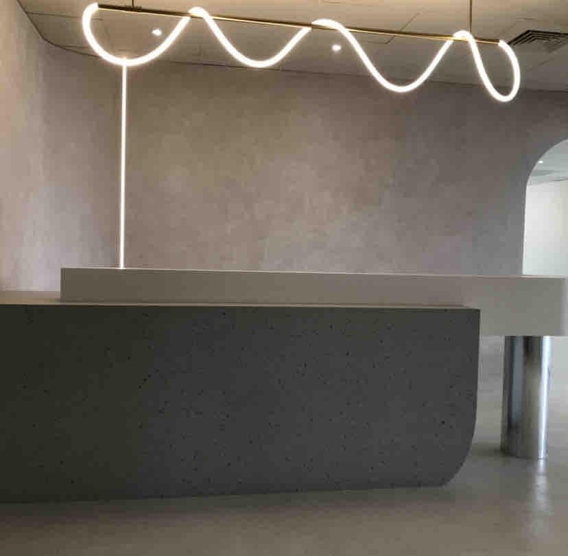 1) 這張照片顯示了一個現代的接待台,接待台是灰色的,並帶有光滑的表面,牆壁是淺灰色的,並帶有白色曲線的燈,燈在牆壁上投下陰影