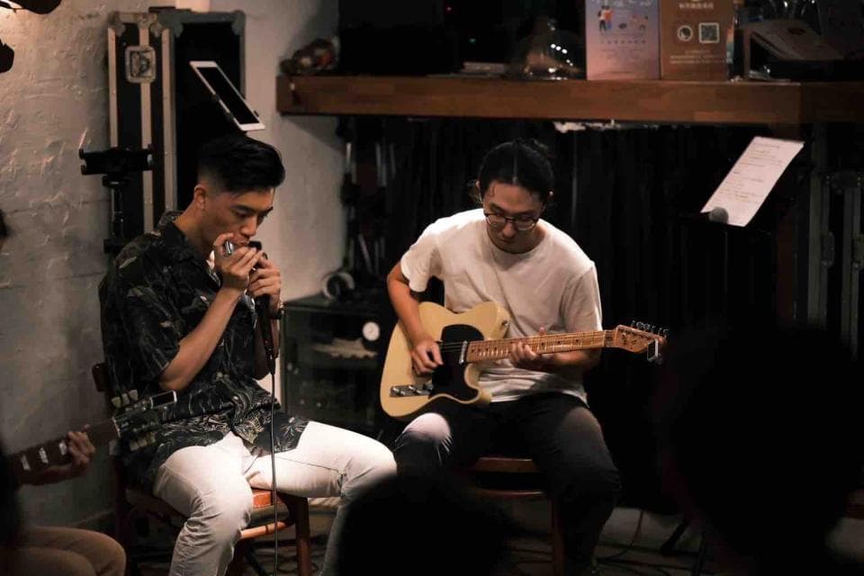 這張「兩個男人在室內練習音樂」的照片是,一個男人坐在椅子上吹口琴,另一個男人坐在椅子上彈吉他。