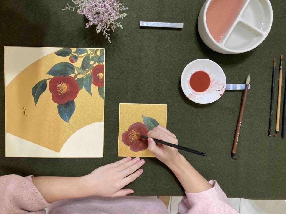 這張照片是一個女性正在用毛筆在紙上畫一朵紅色山茶花。