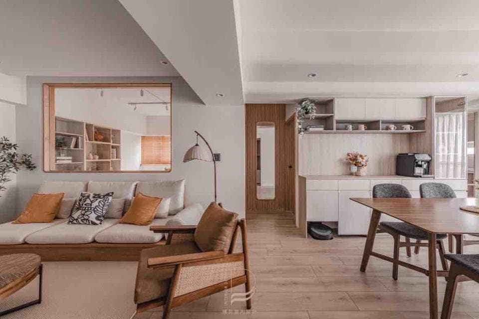 這張「現代簡約風格」的照片是一個客廳,以淺色木質家具和白色牆壁為主。客廳裡有一個大型的沙發、一張木質茶几和一個帶有藤製座椅的木質扶手椅。牆壁上有一個木製的鏡框,鏡框裡可以看見房間的一部分。客廳的旁邊是廚房區域,可以看到木質餐桌和椅子。整体空间明亮简洁,营造出舒适放松的氛围。