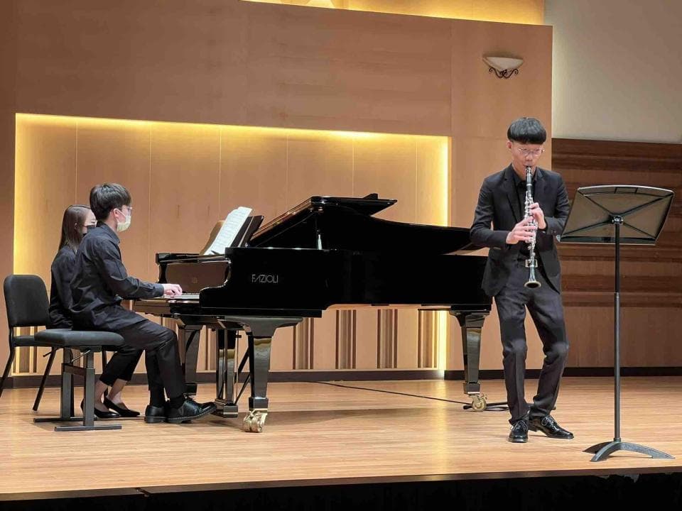 這張照片是一位演奏單簧管的音樂家,他穿着黑色西裝,戴着黑色領帶,拿着單簧管站在舞台上,正準備演奏。