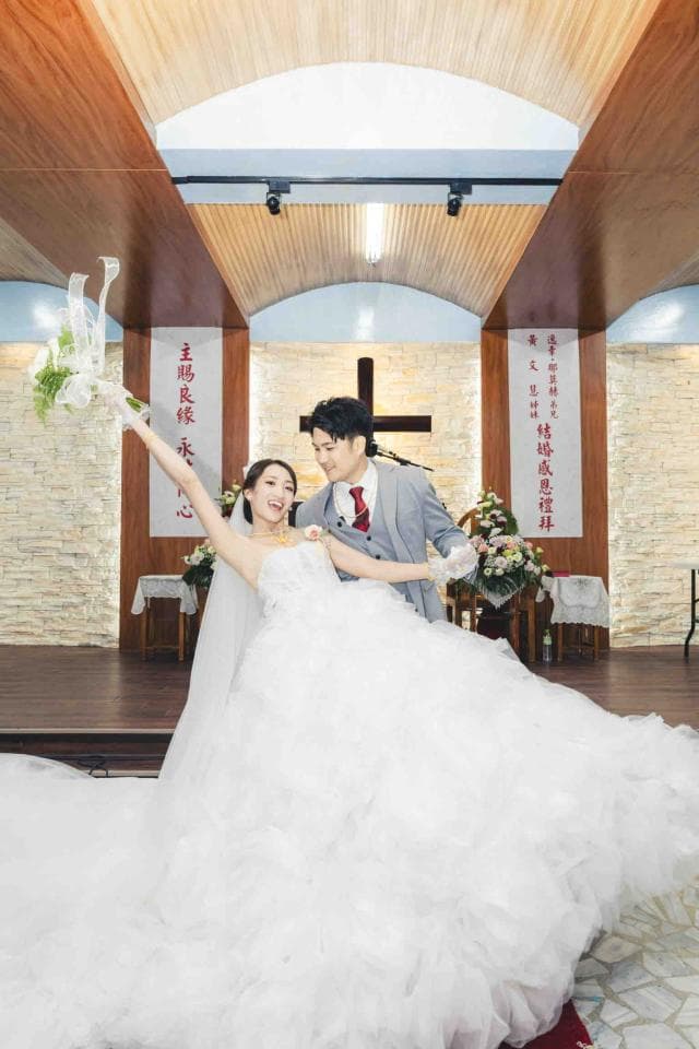 這張照片是一對新人正在教堂舉行婚禮。新娘穿著蓬鬆的白色婚紗,新郎穿著灰色西裝,兩人開心地手牽手,在眾人見證下,邁向人生新的階段。