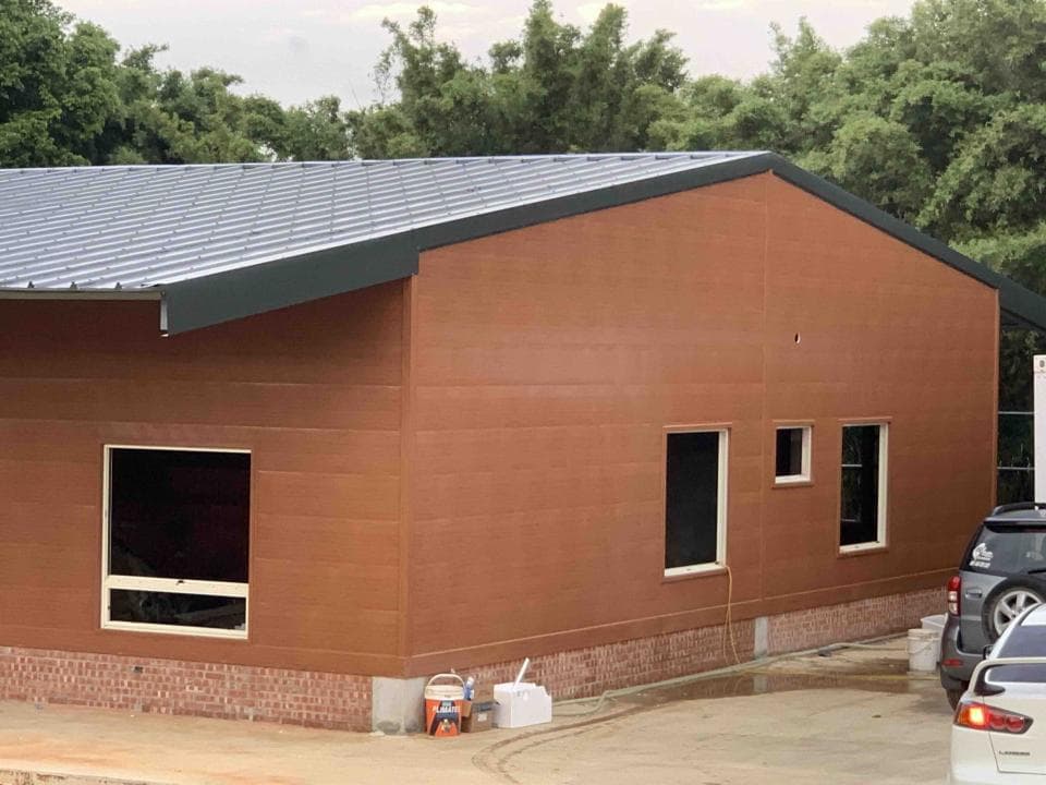 這張照片是一棟正在施工的建築,外牆以紅褐色的牆板包覆,屋頂則以灰色金屬板覆蓋。建築的正面有幾扇窗户,窗户旁放置着一些白色塑膠桶,可能是用來裝水或施工材料的。