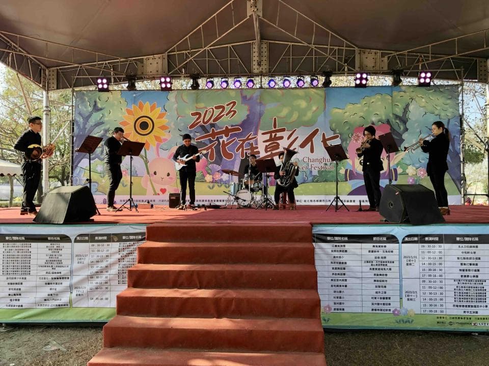 這張照片是一場戶外音樂表演,舞台上有一群樂手正在演奏,背景是綠色的樹木和花朵,舞台前方的階梯上鋪著紅色地毯,整體氛圍歡快熱鬧。