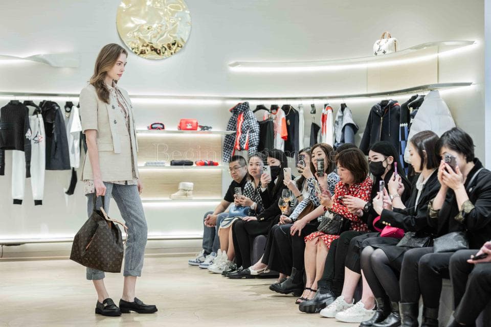 這張「時尚活動」的照片是,一個穿著米色外套和牛仔褲的女性模特正在展示一個大型棕色皮革手提包,她後面是一群身穿時尚服裝的人,他們正在觀看模特。