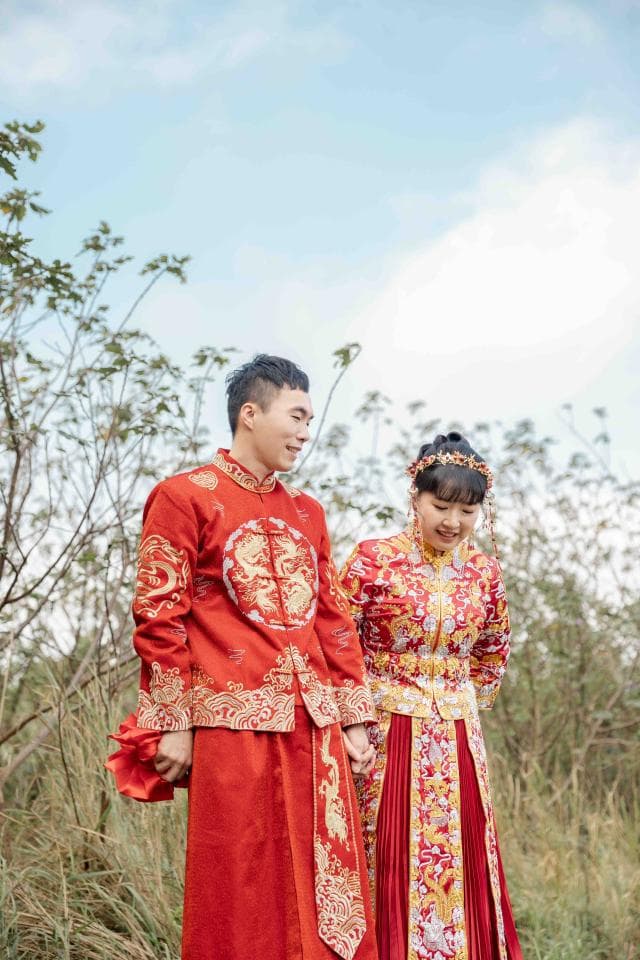 這張照片是一對身穿傳統中式禮服的新人,他們手牽著手,站在戶外,背景是綠色的植物。