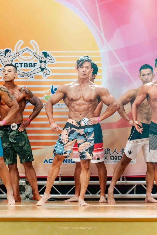 這張照片是一個健美比賽的場景,最重要的是中間穿著花色泳褲的健美選手。他的肌肉線條明顯,表情自信,展現出精壯的身材。