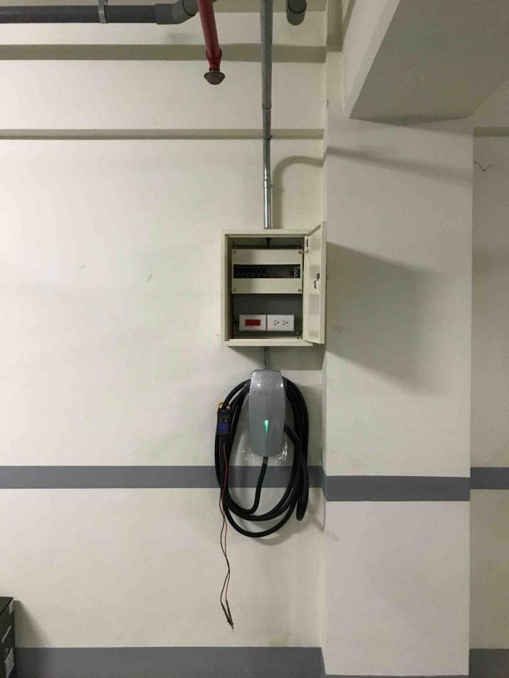 這張照片是一個牆壁上的電動汽車充電站。充電站是銀色的,有黑色電線連接。充電站下方有一個白色電線盒,裡面有開關。