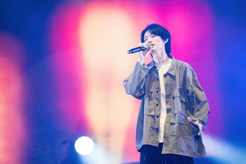 這張「演唱會」的照片是一個歌手在臺上唱歌,他穿著一件灰色外套,拿著麥克風,背景是模糊的燈光。