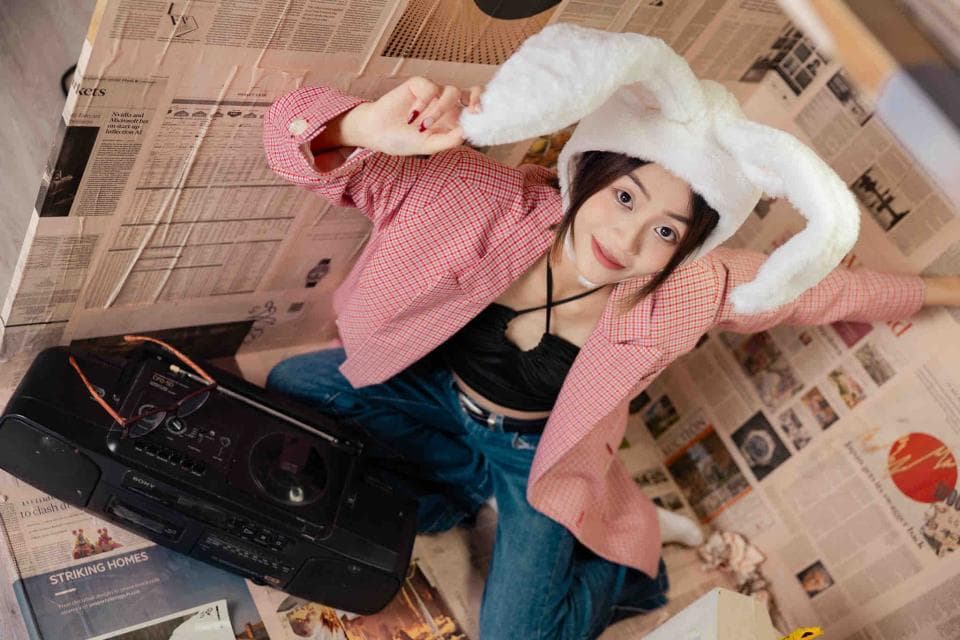 這張照片是一個女生戴著毛茸茸的兔耳朵帽子,她穿著粉紅色的外套,坐在一堆報紙上。