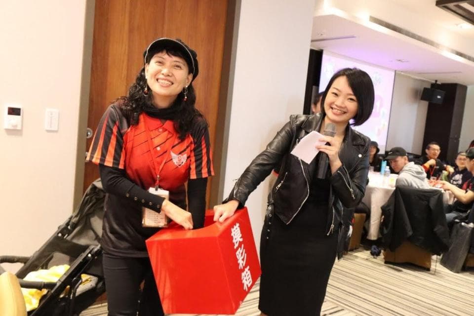 這張照片是兩個女人站在一起,其中一個女人拿著麥克風,而另一個女人正在紅色捐款箱放錢。