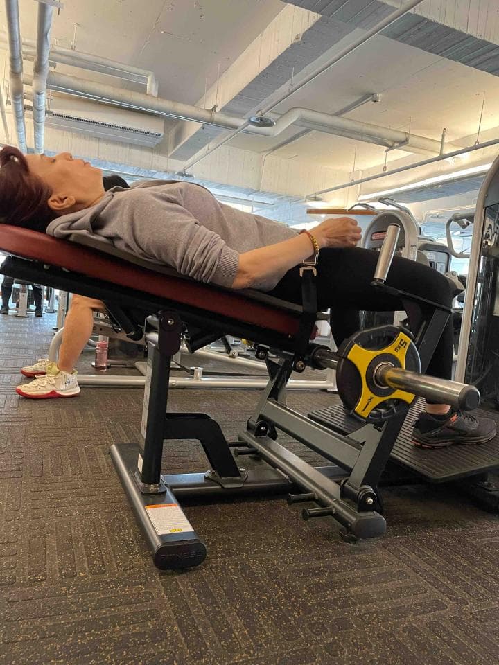 這張照片是一名女子正在健身房使用腿部訓練器材。她躺在器材上,雙腿伸直,並透過器材的力量來鍛鍊腿部肌肉。
