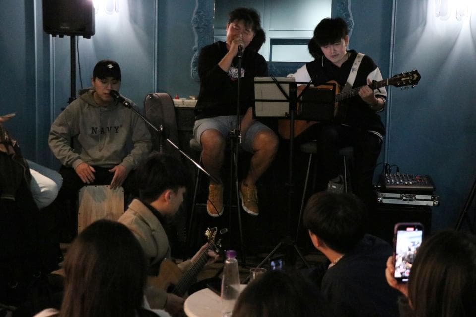 這張照片是一個在酒吧裡演奏音樂的樂隊。樂隊裡有一位正在彈吉他,另一位正在唱歌,還有一位正在打鼓。他們正在演奏一首歡快的歌曲,臺下的觀眾們都在享受音樂。