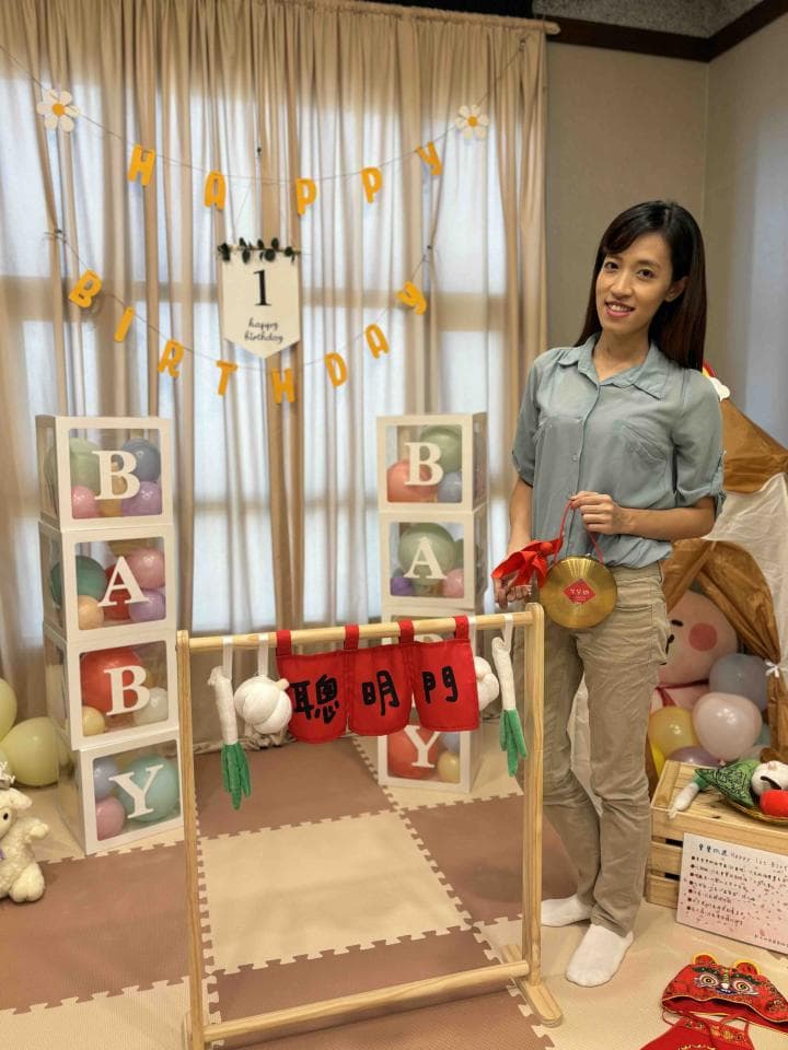 這張「寶寶週歲派對」的照片是,一個女孩站在一個裝飾著彩色氣球和「HAPPY BIRTHDAY」字樣的房間裡,背景佈置著許多氣球和一個裝飾著「1」的牌子。