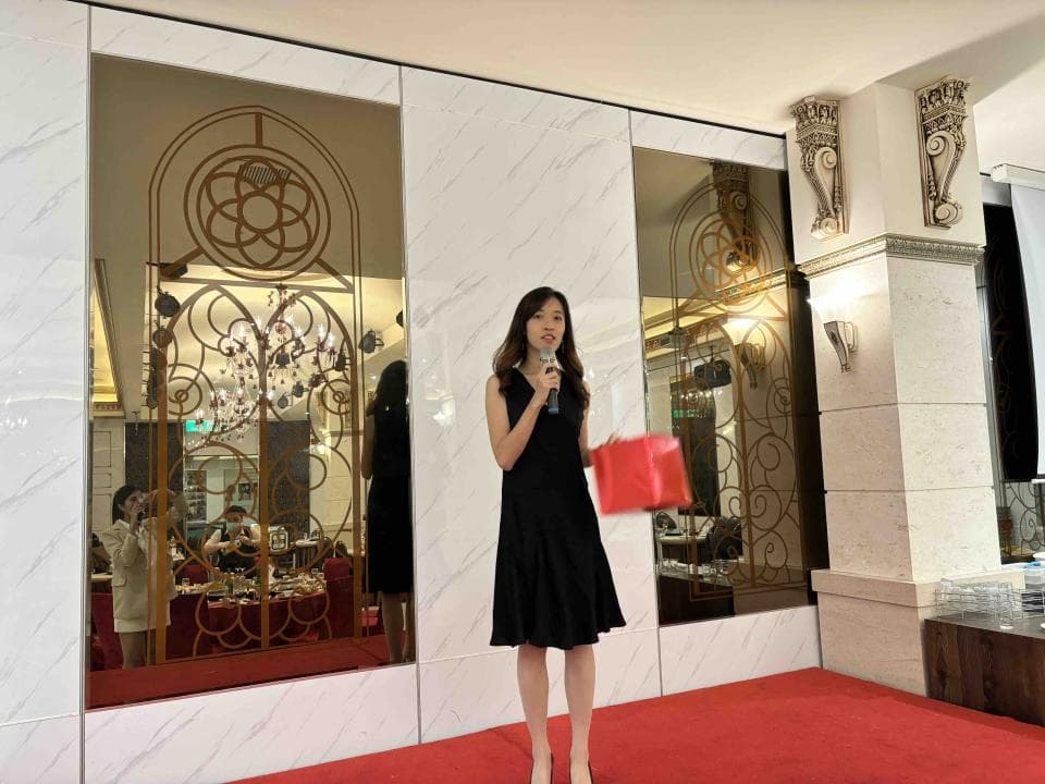 這張「一位女士站在舞臺上發表演講」的照片是,她穿著黑色洋裝,手持紅色禮物盒,並拿著麥克風說話。舞臺上有紅色地毯,背景是裝飾華麗的鏡面牆。