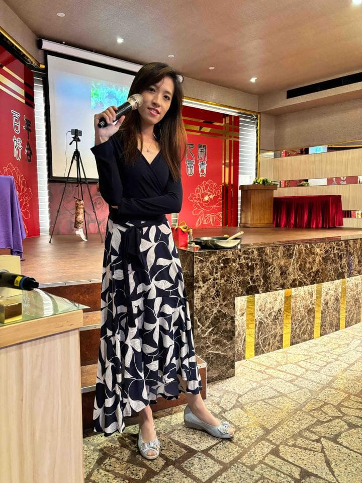 這張照片是一名女子站在舞臺上,她穿著黑色上衣和白色花紋裙,拿著麥克風,看起來像是準備唱歌。