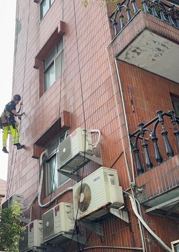 1) 照片顯示一名身穿安全服的工作人員,使用繩索攀爬在建築物的牆壁上,建築物的牆壁上有許多空調室外機
