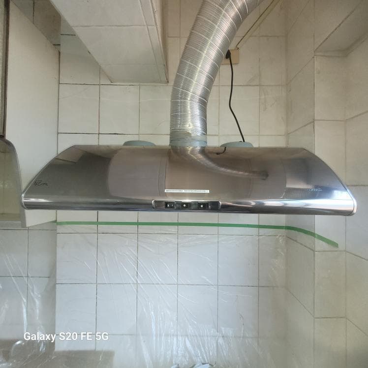 這張照片是一個現代化的廚房,照片中最重要的部分是安裝在牆上的抽油煙機。抽油煙機的材質是銀色的不鏽鋼,設計簡潔,整體看起來非常乾淨,而且它還有排風管連接到屋頂,可以有效地吸走油煙。