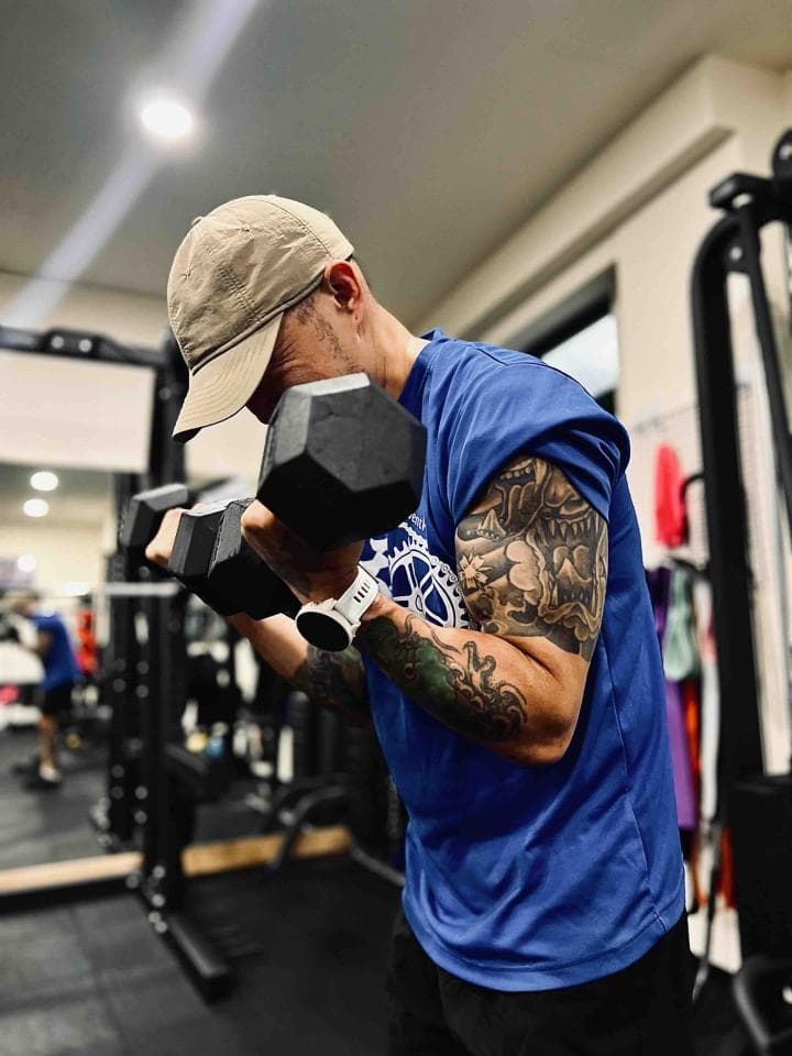 這張照片是一個男人正在健身房舉重。他穿著藍色的運動上衣,右手戴著手錶,正在舉起黑色的啞鈴。