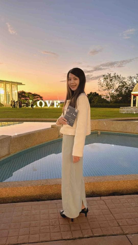 這張照片是一個穿著白色外套的女孩站在一個池塘邊,她拿著一本書,背景是夕陽。