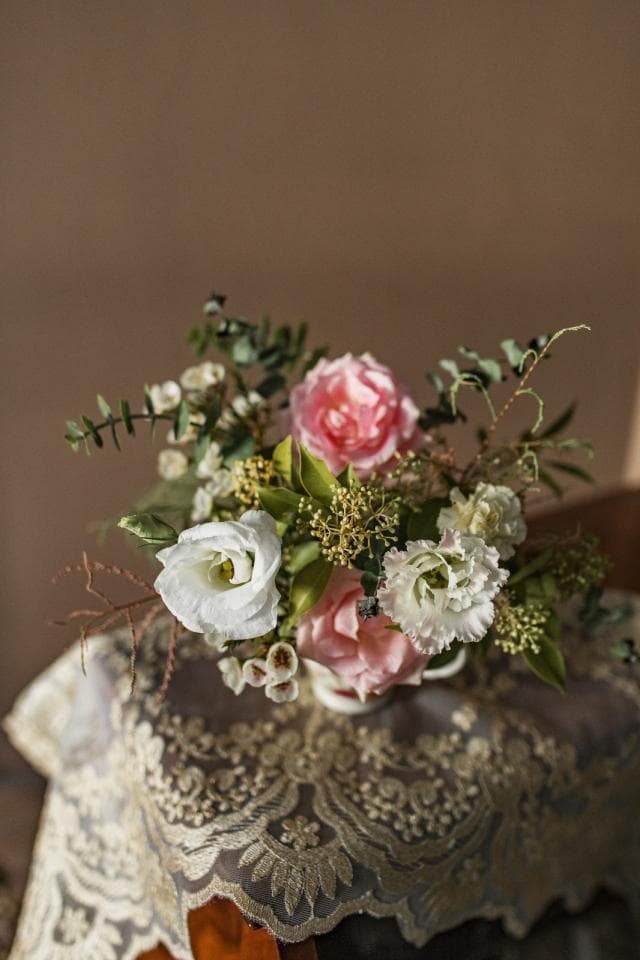 1) 照片中是一束粉紅色和白色的花,花束放在蕾絲桌巾上,背景是土褐色的牆壁