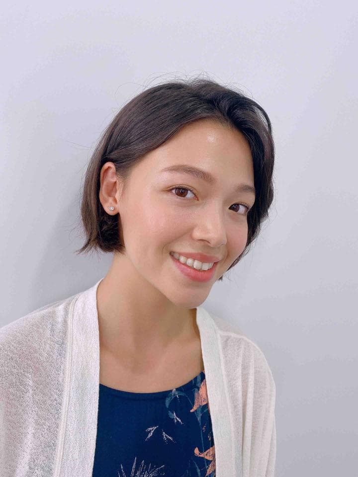 這張照片是一個年輕女性的頭像。她穿著一件白色外套,露出黑色印花背心,短髮,笑得很開心。