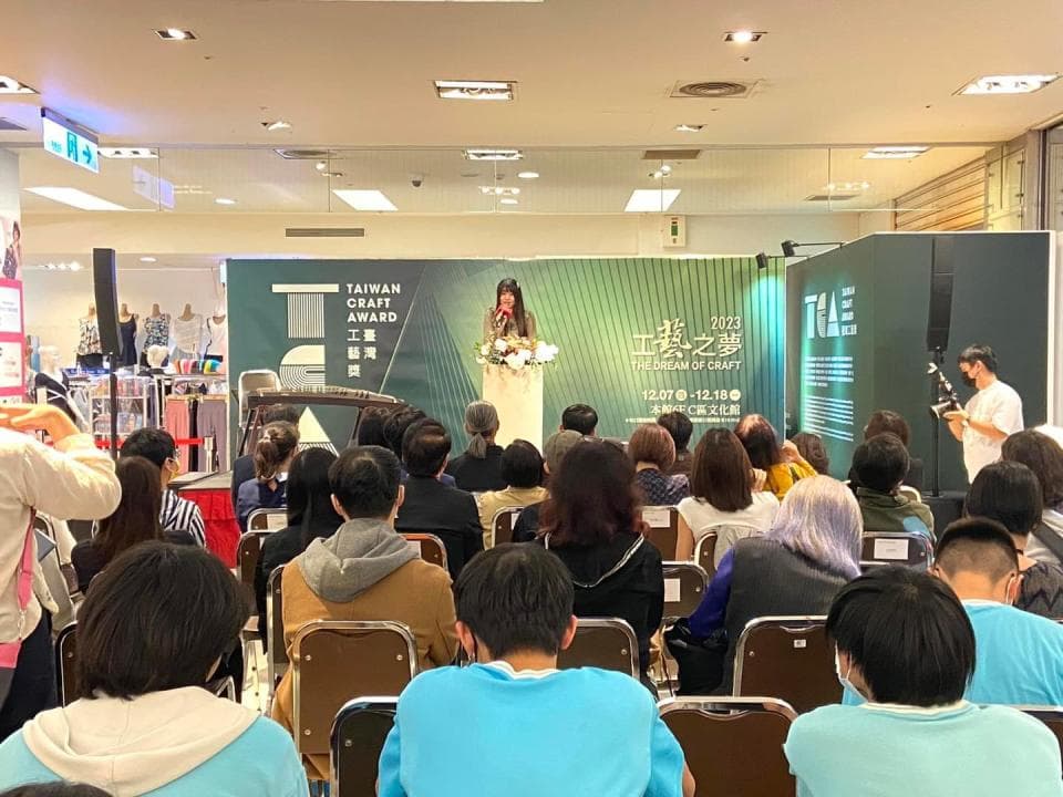 這張照片是 2023 年臺灣工藝之夢活動現場,臺上有一位女性正在致詞,臺下有許多觀眾聆聽。