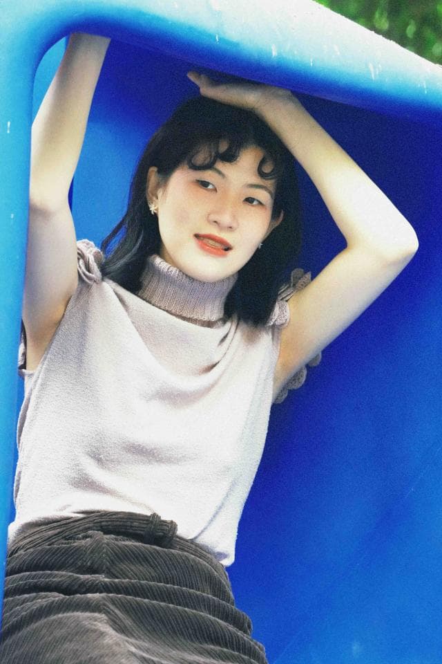 這張「女孩在藍色背景下擺姿勢」的照片是,她穿著淺粉色毛衣和深色裙子,一隻手放在頭頂,另一隻手放在身體兩側,面帶微笑。