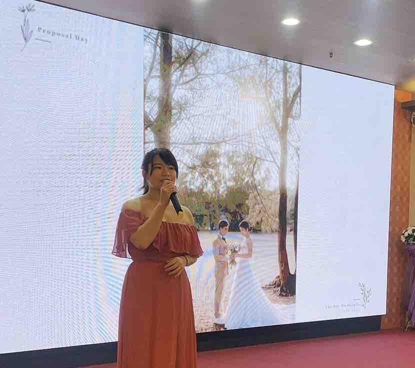 這張「婚禮」的照片是一名穿著紅色禮服的女性站在大螢幕前,她手持麥克風,而螢幕上播放著一對新人在戶外婚禮的照片。