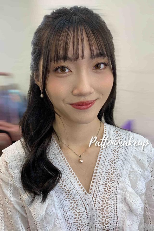 這張照片是一個年輕女子,她穿著一件白色蕾絲上衣,配戴一條金色的項鍊,項鍊上有一個小巧的珍珠吊墜。她擁有烏黑亮麗的長髮,留著齊瀏海,散發著溫柔甜美的氣質。