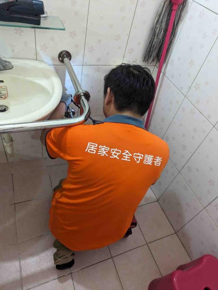 1) 照片顯示一位身穿橘色上衣、上面印有「居家安全守護者」字樣的男性,正在浴室修理洗手台